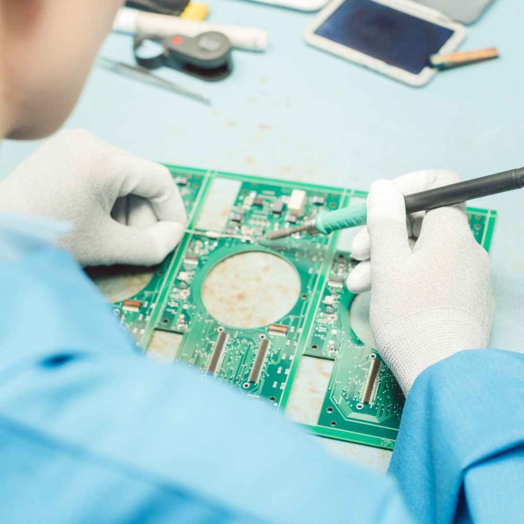 Image of a technician manually soldering components onto a printed circuit board (PCB) with a soldering iron. The technician is wearing gloves and a lab coat, indicating a controlled assembly environment. This image showcases Bencor's skilled manual assembly for precise PCB work.